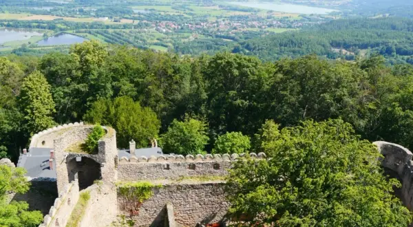 Kamienne mury zamku Chojnik wznoszą się wśród zieleni, a w oddali rozciąga się malowniczy krajobraz z jeziorami i miasteczkiem.