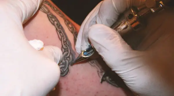 Manos enguantadas tatuando una cadena rota en la piel.