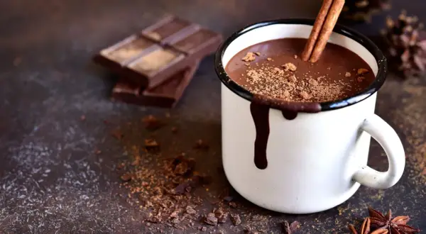 Cioccolata calda cremosa con cacao e cioccolato, guarnita con cannella e cacao in polvere. Accanto, pezzi di cioccolato fondente e spezie.