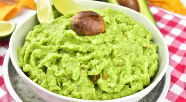 Miseczka guacamole z pestką, limonką i nachos w tle na czerwono-białej kratce.