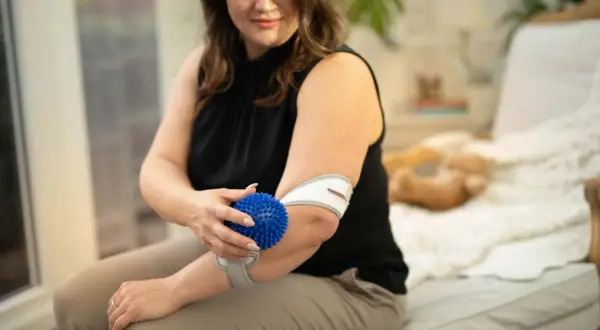 Frau massiert ihren Unterarm mit einem blauen Igelball und trägt eine Bandage.