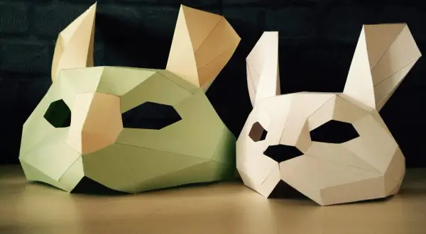 Två påskpyssel i papper: en ljusgrön och en vit kaninmask med stora öron och geometriska former.