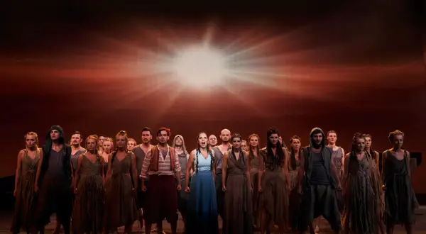 Chór w "Aidzie" w Teatrze Roma, spektakl trwa. Grupa ludzi w strojach z epoki na tle dramatycznego, rozświetlonego nieba.