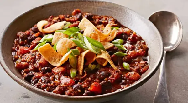 En skål med mustig, lakto-ovo vegetarisk chili med bönor, toppad med krispiga tortillachips och salladslök. En sked ligger bredvid.