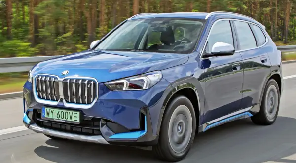 Niebieski BMW X1 elektryk pędzi po drodze wśród drzew.