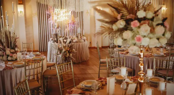 Elegancka sala weselna z neonem "All you need is love". Złote krzesła, pudrowo-różowe obrusy i bujne kwiaty. Zastanawiasz się, ile kosztuje udekorowanie sali weselnej w takim stylu?