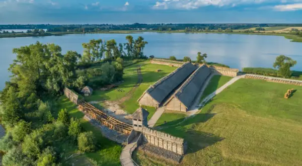 Rekonstrukcja osady Biskupin z drewnianymi domami krytymi strzechą, wałami obronnymi i jeziorem w tle.