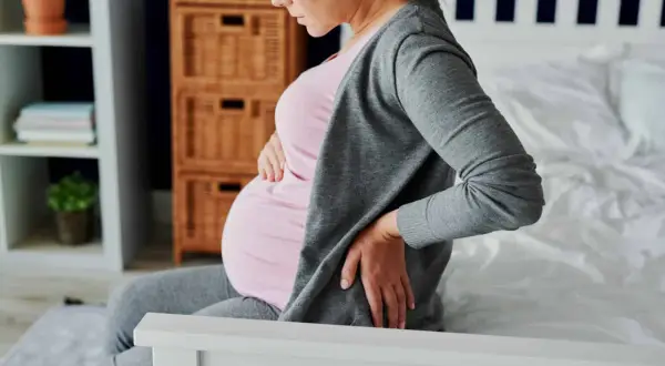 Femme enceinte assise sur un lit, tenant son ventre et se tenant le bas du dos, souffrant peut-être d'hémorroïdes.