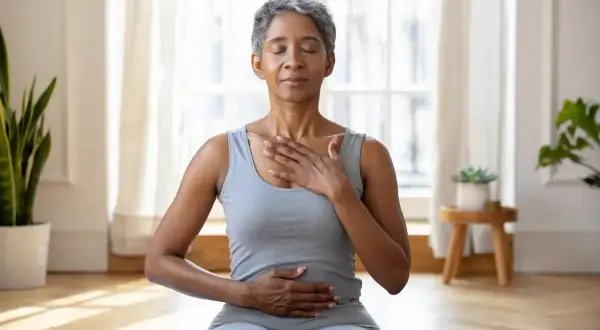 Femme pratiquant une technique de relaxation mentale, mains sur la poitrine et le ventre, yeux fermés, dans une pièce lumineuse.
