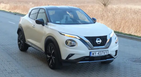 Biały Nissan Juke Sport z czarnym dachem i felgami, zaparkowany na kostce brukowej przy suchej trawie.