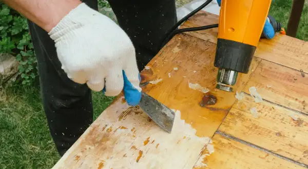 Main tenant un décapeur thermique et une spatule pour retirer la colle sur bois vernis.