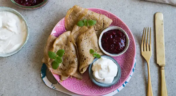 En tallrik med glutenfria pannkakor, serverade med sylt och grädde. En gyllene gaffel och kniv ligger bredvid.