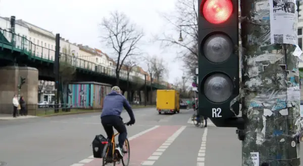 Ein Radfahrer wartet an einer roten Ampel. Im Hintergrund eine gelbe LKW und eine Hochbahn.