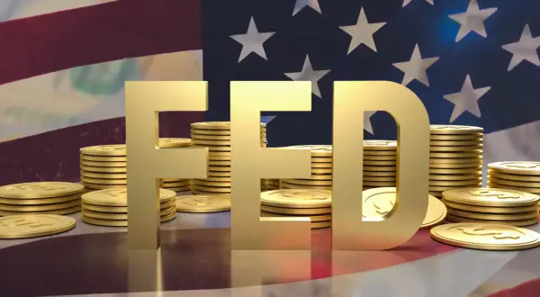 Złote litery "FED" na tle flagi USA i stosów monet. FOMC to komitet, który decyduje o polityce pieniężnej.