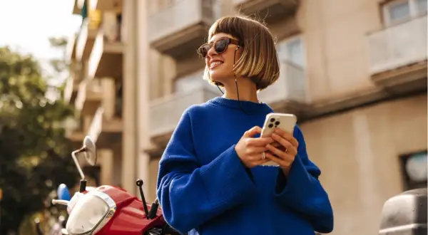 Frau mit blondem Bob, Sonnenbrille und blauem Pullover, die auf ihrem Handy scrollt. Ein rotes Moped steht daneben. Ein toller Look, wie man einen blauen Pullover kombinieren kann, Herren-Style inspiriert.