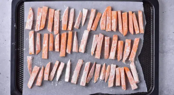 Pokrojone frytki z batatów posypane mąką na blasze do pieczenia.