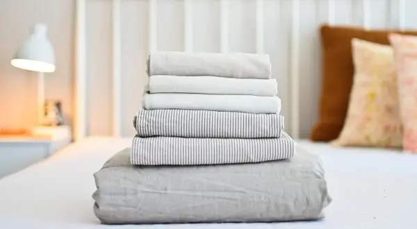 Neatly folded stack of clean bed sheets on a white bed, ready for how to wash bed sheets in washing machine.