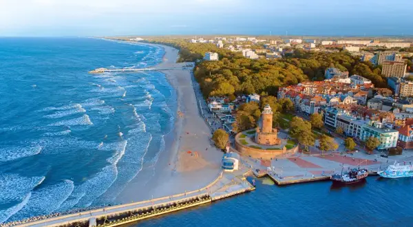 Widok z lotu ptaka na **latarnię morską Kołobrzeg** i wybrzeże Bałtyku. Widać plażę, molo i miasto.