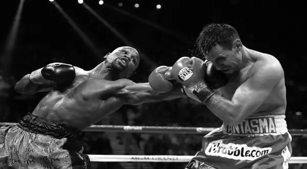 A boxer throws a powerful double jab, sweat flying as his opponent braces for impact in the intense spotlight of the ring.