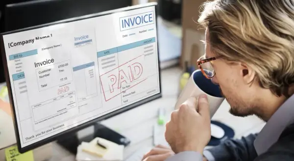 Mężczyzna pije kawę, patrząc na ekran komputera z fakturą z pieczątką "PAID" i informacją o podatku VAT.