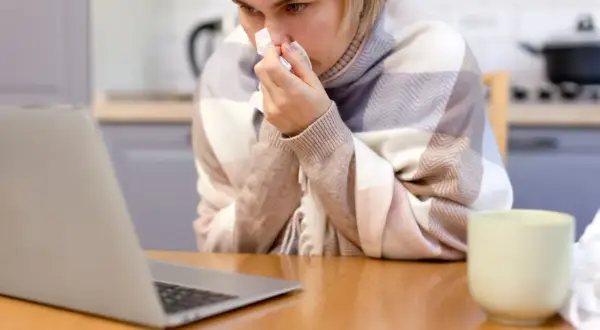 Kobieta w kratę, z chusteczką w dłoni, korzysta z laptopa. Może to być polskie zwolnienie lekarskie w Danii, gdy czuje się źle.