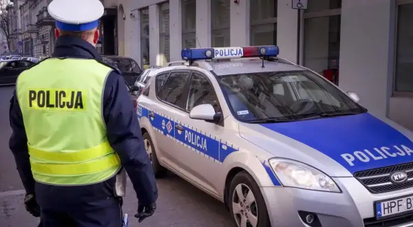 Policjant w kamizelce odblaskowej stoi obok radiowozu z napisem "POLICJA".