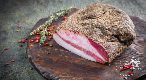 Pancetta co to? Wędzony kawałek boczku z widocznymi warstwami tłuszczu i mięsa, przyprawiony pieprzem, leży na drewnianej desce z ziołami i ziarnami pieprzu.