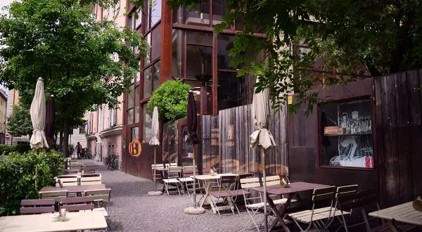 Tavoli all'aperto e ombrelloni di fronte al Batzen Bräu, con un'architettura industriale e un'atmosfera rilassata.