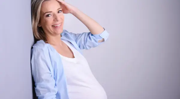Uśmiechnięta kobieta w ciąży po 40. opiera się o ścianę, dotykając włosów.