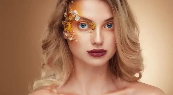 Fotografia beauty: kobieta z niebieskimi oczami, ozdobiona kwiatami i złotym makijażem na twarzy.
