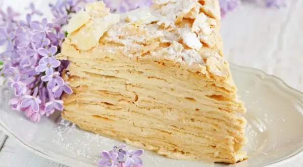Pyszny tort napoleon z delikatnym kremem i chrupiącymi warstwami ciasta, ozdobiony płatkami migdałów i cukrem pudrem, w otoczeniu kwitnącego bzu.