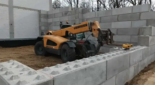 Żółty ładowacz teleskopowy Liebherr układa betonowe bloki, budując mur oporowy.