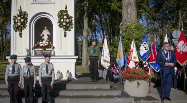 Uroczystość przy kapliczce. Czy imię Bruno jest święte? Dzieci w harcerskich mundurkach i dorosły z łańcuchem na szyi stoją przed kapliczką z figurą i flagami.