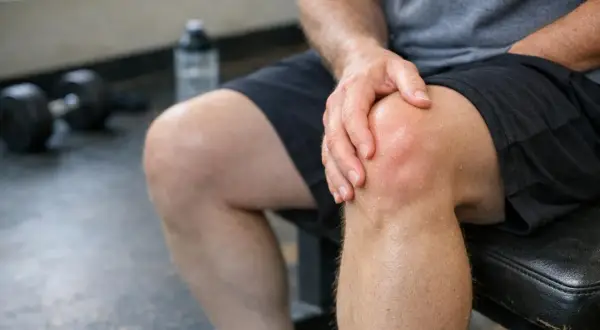 Uomo con mano sul ginocchio, evidenziando il gonfiore post allenamento. Panca e pesi sullo sfondo.