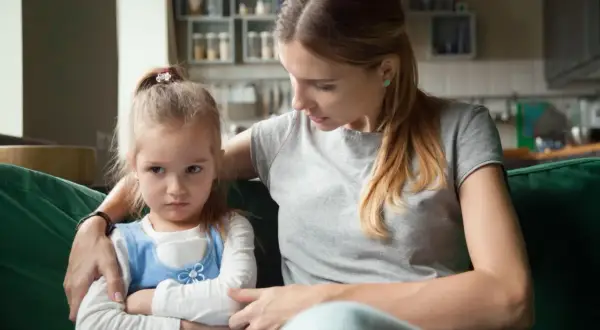 Mama próbuje oswoić złość swojej córki, która siedzi obrażona z założonymi rękami.
