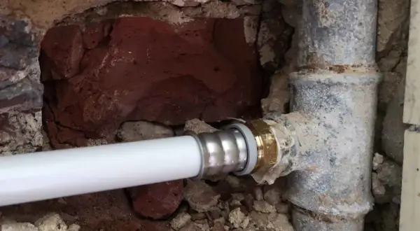 Un nouveau tuyau blanc est raccordé à un ancien tuyau de plomb pour l'eau. La maçonnerie est visible autour des canalisations.