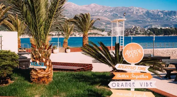 Palmy, morze i góry w Czarnogórze. Znaki wskazują plażę, Aperol Spritz i "Orange Vibe". Idealne miejsce na wakacje.