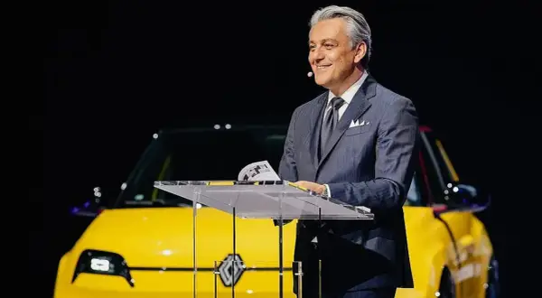 Hombre sonriente en traje, presentando un coche amarillo eléctrico, quizás un nuevo modelo de Volvo Renault.