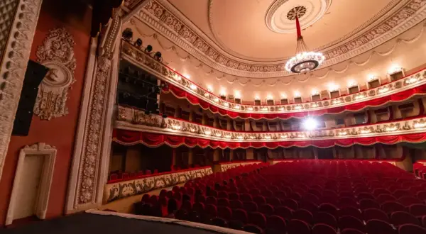 Wnętrze teatru z czerwonymi fotelami i bogato zdobionymi lożami. Czekamy na rozpoczęcie spektaklu, by usłyszeć piękną **arię**.