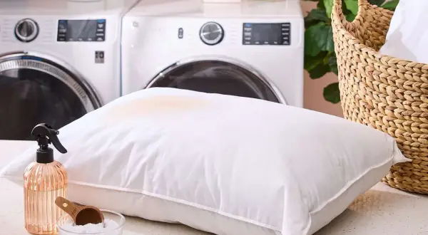 Learn how to get smell out of pillows with this guide. A stained pillow, spray bottle, and baking soda are shown, with laundry machines in the background.