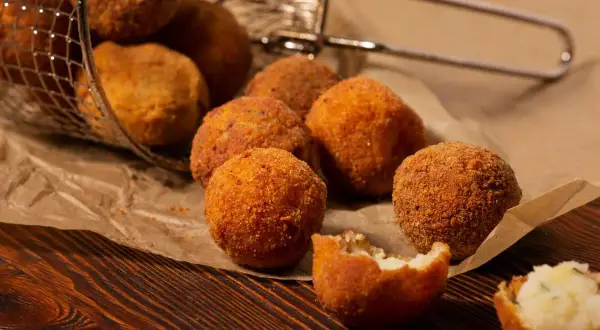 Croquetas de patata y pollo doradas y crujientes, algunas en una cesta metálica, otras sobre papel de estraza.