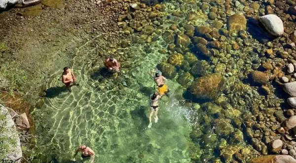 Gente se baña en un río cristalino con fondo de piedras, cerca de un **ponton de la oliva**.