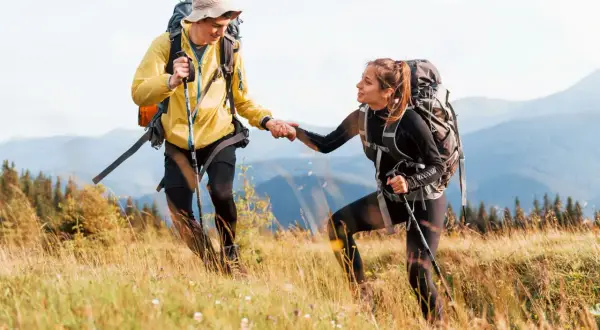 Coppia di escursionisti in montagna si aiutano a salire un pendio erboso, con zaini e bastoncini da trekking.