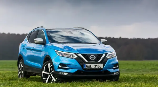 Niebieski Nissan Qashqai 1.6 benzyna na tle zielonej łąki i pochmurnego nieba.