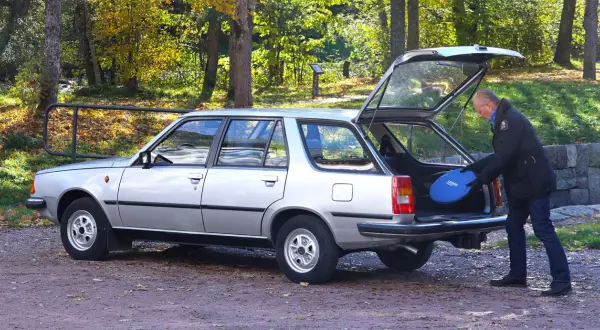 Man lastar en blå frisbee i bakluckan på en silverfärgad Renault 18 kombi i höstlandskap.