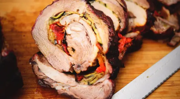 Coppa di maiale allo spiedo, succulenta e ripiena di verdure, pronta per essere affettata su un tagliere di legno.