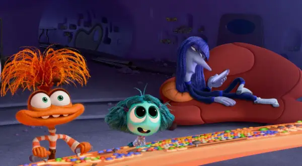 Hier ist alles steht kopf 2 emotionen: Ein orangefarbenes Wesen mit struppigen Haaren drückt Knöpfe, ein kleines blaues Wesen schaut neugierig, während ein langes, blaues Wesen auf einer Couch liegt und auf sein Handy schaut.