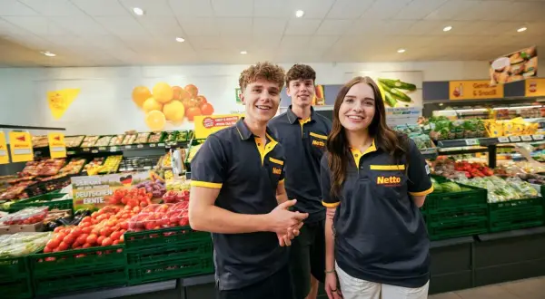Drei junge Mitarbeiter lächeln in einem Supermarkt. Sie tragen Arbeitskleidung mit dem Netto-Logo. Die Ausbildung zum Elektroniker bietet ein gutes Gehalt netto.