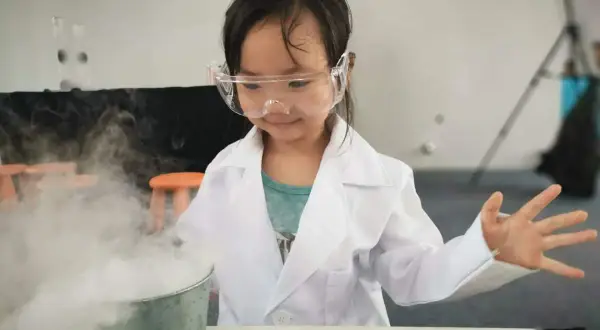 Niña con bata y gafas realiza experimentos para niños de 7 años, creando humo blanco de un cubo.