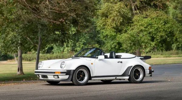 Biały Porsche 911 Speedster z lat 80. z czarnym dachem i spojlerem, zaparkowany na tle drzew.